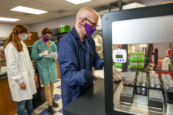 Rachel Ham, left, Clemson University lab supervisor, lets  Dylan Chitwood, a Clemson University Bioengineering PhD student, look over paperwork while Jonathan Heywood, right, a Clemson University PhD student, works with a robot processing saliva in the COVID-19 Clinical Diagnostics Lab at Clemson University.